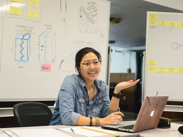 A student sits at a desk in front of a pair of whiteboards with sketches on them. She has her laptop computer open and appears to be speaking with someone off-camera.